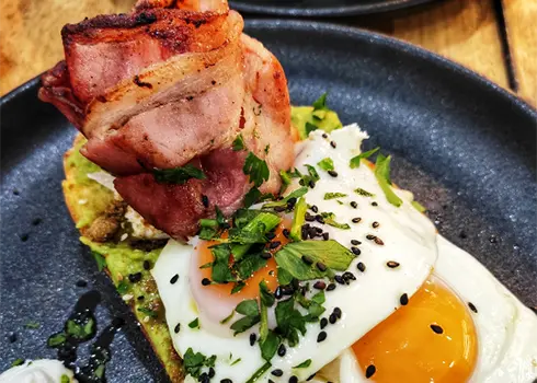 Close Up Of A Plate Of Smashed Avocado Toast, Topped With Bacon & Fried Eggs